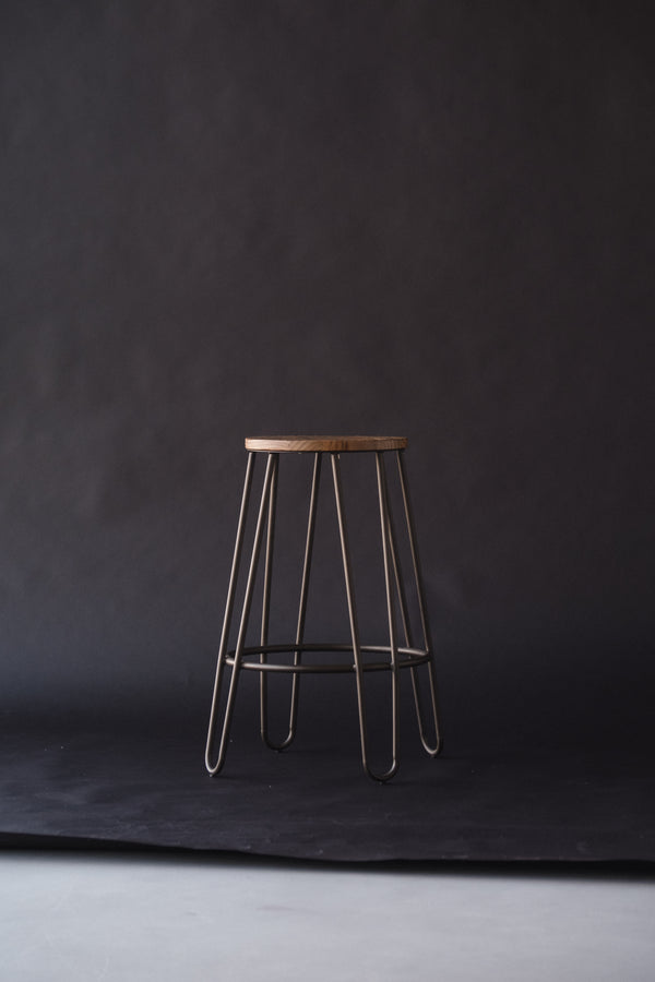 A wooden stool with hairpin legs against a dark studio backdrop